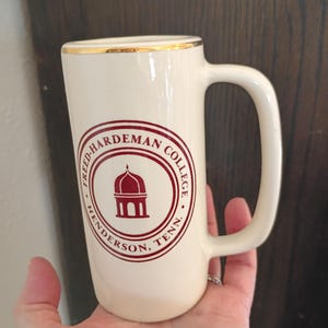 Puede incluir: Taza de cerámica color crema con borde dorado y asa grande. La taza presenta un logotipo circular granate con el texto "FRED-HARDEMAN COLLEGE HENDERSON, TENN." y una ilustración estilizada de un edificio.