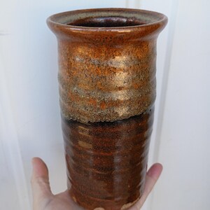 Puede incluir: Un jarrón de cerámica hecho a mano con forma cilíndrica. El jarrón presenta una superficie texturizada con un degradado de tonos marrones y bronce. El borde superior es ligeramente acampanado, y el jarrón se sostiene en una mano.