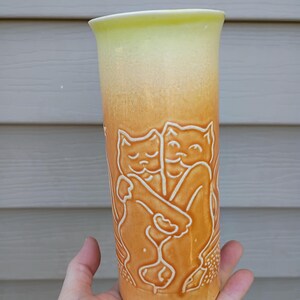 May include: A tall, ceramic vase with a gradient of yellow to orange hues. The vase features an embossed design of two embracing cats. The cats are in a light cream color, contrasting against the orange background. The vase is held in a hand.