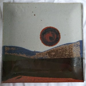 May include: A ceramic plate with a landscape scene painted on it. The scene features a large red sun setting behind a blue and brown mountain range. The plate has a white background with brown speckles.