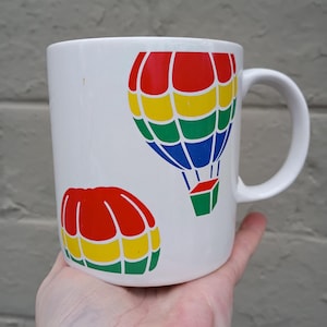 May include: White ceramic mug featuring two colorful hot air balloon designs. The balloons are in red, yellow, green, and blue. The mug has a curved handle and is held in a hand.