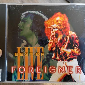 Könnte beinhalten: CD-Hülle für das Album "Live Best of Foreigner". Das Cover zeigt ein Live-Performance-Bild eines Sängers mit roten Haaren und Mikrofon, mit einem grün getönten Bild eines anderen Sängers im Hintergrund. Der Albumtitel ist in Orange und Weiß gehalten.