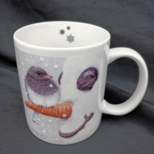Può includere: Tazza in ceramica bianca con una scena invernale, con un uccello posato su un ramo di carota. La tazza ha un manico ed è decorata con fiocchi di neve e un disegno di pupazzo di neve. L'uccello ha piume grigie e bianche.
