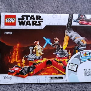 May include: LEGO Star Wars set featuring a scene with two figures wielding blue lightsabers. The set includes a lava-themed background and a mechanical arm. The LEGO logo and Star Wars text are visible, along with the Disney logo.