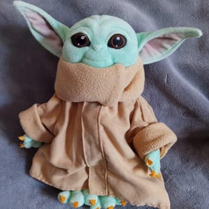 May include: A plush toy of a character with large ears, big eyes, and a tan robe. The character has a light green face and hands, with orange claws on its feet and hands. The toy is on a grey surface.