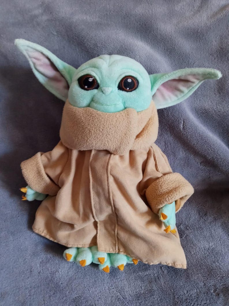 May include: A plush toy of a character with large ears, big eyes, and a tan robe. The character has a light green face and hands, with orange claws on its feet and hands. The toy is on a grey surface.