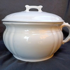 Puede incluir: Orinal de cerámica blanca con tapa y asa. El orinal tiene forma redondeada con estrías verticales y un asa lateral. La tapa tiene un pequeño asa en la parte superior.