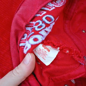 May include: A close-up of a red baseball cap with white and blue embroidered text inside. The cap has a tag that reads "alpha" and lists the materials. The cap is made of 50% cotton and 50% polyester.