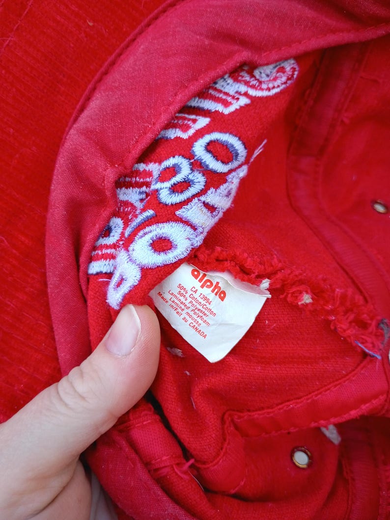 May include: A close-up of a red baseball cap with white and blue embroidered text inside. The cap has a tag that reads "alpha" and lists the materials. The cap is made of 50% cotton and 50% polyester.