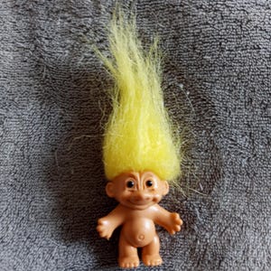 Könnte beinhalten: Eine Vintage-Troll-Puppe mit einem hellbraunen Körper, kleinen Armen und Beinen und einem lächelnden Gesicht. Die Puppe hat ein großes, hellgelbes, stacheliges Haar. Die Puppe ist ein klassisches Spielzeug.