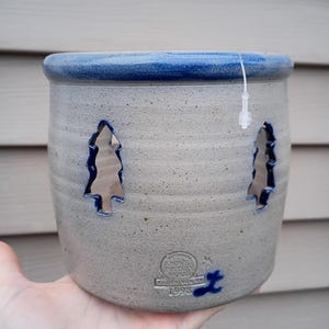 Puede incluir: Un jarrón de cerámica de gres con borde azul y recortes de árboles. El jarrón es de color gris claro con un acento azul alrededor de la parte superior. El jarrón tiene una etiqueta que dice "Rowe Pottery Works Cambridge, WI 1998".