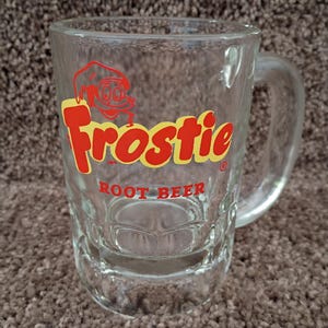 May include: Clear glass mug with a handle, featuring the red and yellow "Frostie Root Beer" logo. The logo includes a cartoon character. The mug has a rectangular shape with a textured base.