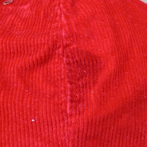May include: Close-up of a bright red corduroy fabric. The fabric has a ribbed texture and a slight sheen. The image shows the detail of the corduroy weave and the color saturation of the material. The fabric appears to be part of a garment.