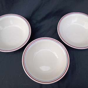 May include: Three white ceramic bowls with a red and blue striped rim. The bowls are empty and have a simple, classic design. The bowls are likely for serving food.