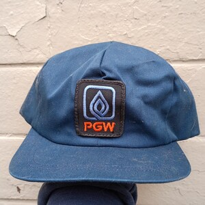 Può includere: Cappellino da baseball blu navy con visiera curva. Il cappellino presenta una toppa rettangolare con un disegno a fiamma e le lettere "PGW" in arancione. La toppa è bordata in nero e blu.