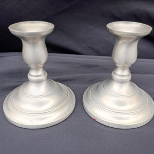 May include: Two silver-colored metal candle holders. Each has a round base, a curved stem, and a shallow cup to hold a candle. The candle holders are displayed on a dark surface.