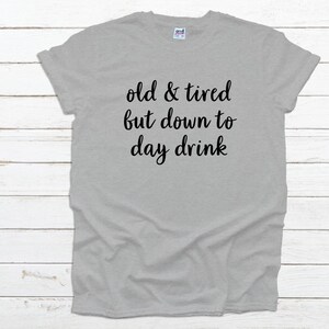 May include: A light gray t-shirt with the text "old & tired but down to day drink" in black script. The shirt is made of a soft material and has short sleeves.