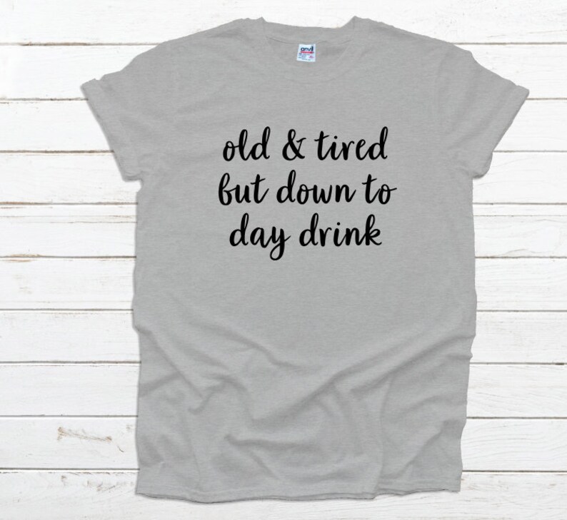 May include: A light gray t-shirt with the text "old & tired but down to day drink" in black script. The shirt is made of a soft material and has short sleeves.