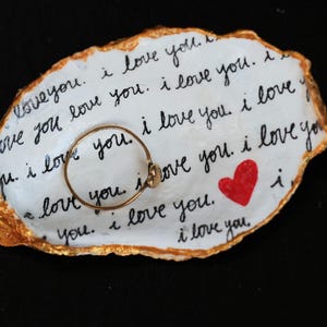 Peut inclure: Un vide-poche ovale blanc avec des bords dorés et l'inscription « I love you » en écriture noire répétée. Une petite bague dorée repose sur le plat, à côté d'un cœur rouge.
