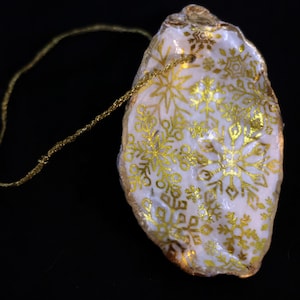 May include: A white oyster shell Christmas ornament with gold snowflake designs and a gold trim. A gold string is attached for hanging. The ornament is set against a black background.