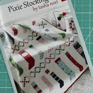 May include: A quilt pattern for a Christmas stocking quilt called "Pixie Stockings" by Tasha Noel. The quilt features a white background with red, green, and blue stockings.