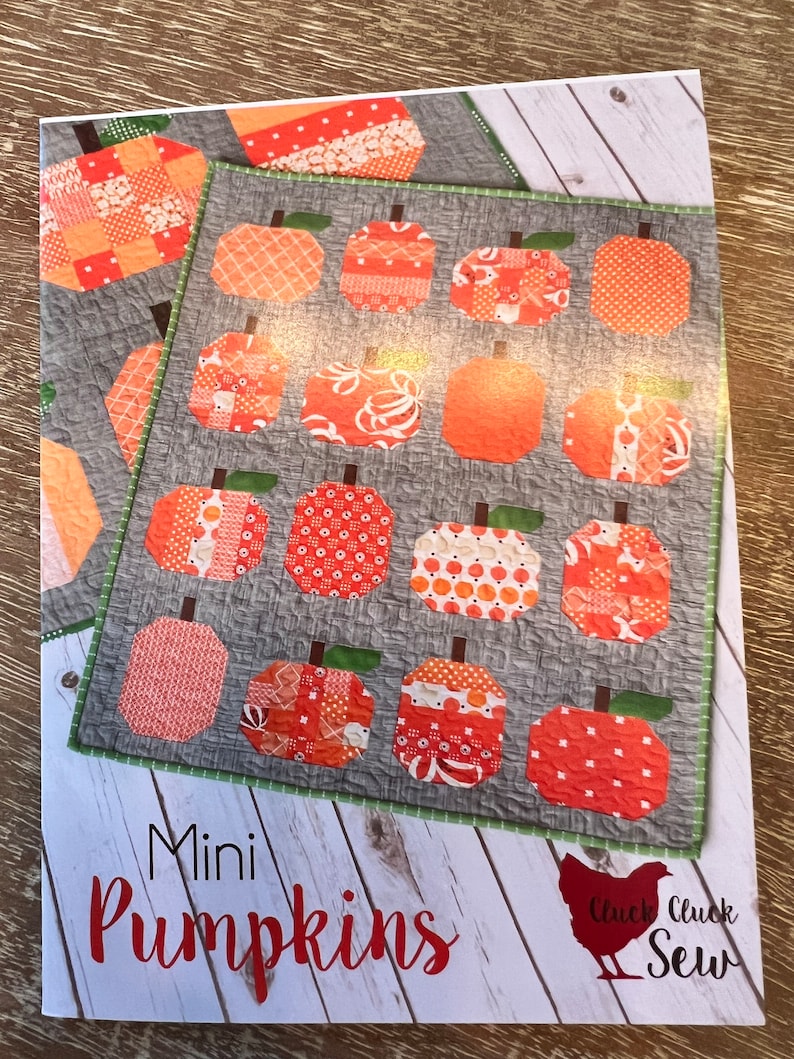 May include: A white and gray printed fabric pattern with a quilt design of orange pumpkins. The text "Mini Pumpkins" and "Cluck Cluck Sew" is printed on the fabric.