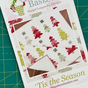 May include: A white fabric quilt with a patchwork design of red and green Christmas trees and stars. The quilt is titled "Tis the Season" and is from The Pattern Basket, Margot Languedoc Designs.