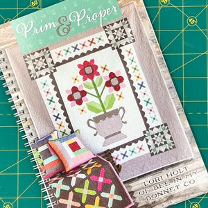 May include: A book titled "Prim & Proper" by Lori Holt of Bee in My Bonnet Co. The cover features a quilt design with a floral motif and a vase. The book is open to a page showing a quilt with a brown background and colourful cross shapes.