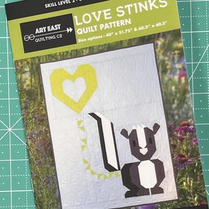 May include: A quilting pattern for a quilt titled "Love Stinks". The pattern features a cartoon skunk with a yellow heart balloon. The pattern is available in two sizes: 40" x 51.75" and 60.5" x 60.5".