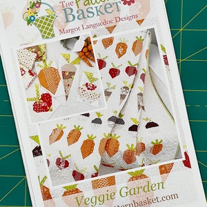 Puede incluir: Un folleto de patrones de papel blanco con una ilustración colorida de una colcha con zanahorias y fresas. El título del patrón es "Veggie Garden" y el texto "The Pattern Basket Margot Languedoc Designs" está en la parte superior de la página. La dirección del sitio web "www.thepatternbasket.com" está en la parte inferior de la página.