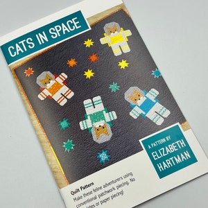 以下が含まれることがあります： エリザベス・ハートマンによる「宇宙の猫たち」というタイトルのキルトパターンの小冊子。表紙は、宇宙飛行士の服を着た4匹の猫と、さまざまな色の星が描かれた黒い背景です。