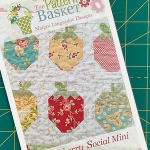 Puede incluir: Un patrón impreso para un edredón titulado "Strawberry Social Mini" de The Pattern Basket. El diseño del edredón presenta seis formas de fresa con varios patrones de tela floral. El fondo es de color crema claro con una textura acolchada.