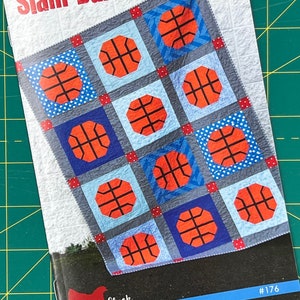May include: A quilting pattern for a quilt featuring a basketball design. The pattern is called "Slam Dunk" and is from Cluck Cluck Sew. The quilt is made with blue, red, and white fabric.