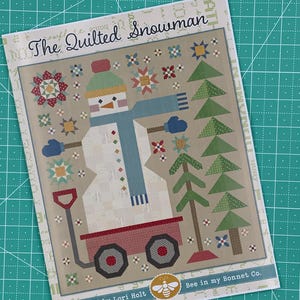 May include: A quilting pattern featuring a snowman in a red wagon with a blue scarf and mittens. The pattern is titled "The Quilted Snowman" and is designed by Lori Holt for Bee in my Bonnet Co.