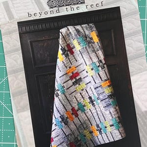 May include: A quilt pattern design with the text "beyond the reef floats". The quilt is made with a variety of fabrics in shades of gray, white, red, orange, yellow, green, and blue. The quilt is folded and draped over a dark wooden surface.