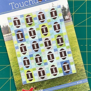 May include: A quilting pattern featuring a football design. The pattern is called "Touchdown" and is from Cluck Cluck Sew. The pattern is number 143.