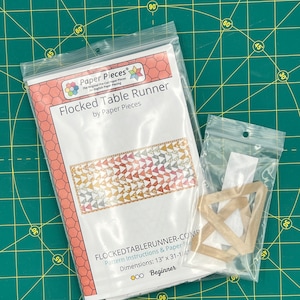 May include: A clear plastic bag contains a "Flocked Table Runner" pattern by Paper Pieces. The pattern instructions are visible, with dimensions of 13" x 31-1/4". A smaller bag holds wooden shapes. The background is a green cutting mat.