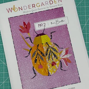 May include: A quilt pattern featuring a colorful, stylized beetle with a yellow body, black and white wings, and red and white flowers. The pattern is titled "The Beastie" and is part of the Wondergarden collection of modern quilt patterns by Tamara Kate. The text "Always be on the lookout for the presence of wonder" ~ E.B. White is also included.