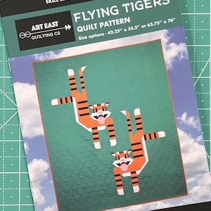 May include: A quilt pattern featuring two orange and black tigers with white stripes, flying upside down against a teal blue background with white clouds. The pattern is titled "Flying Tigers Quilt Pattern" and includes the text "Art East Quilting Co." and "#flyingtigersquilt".