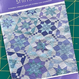 以下が含まれることがあります： 「shimmer」というタイトルのキルトパターンで、モダンで虹色のデザインです。表紙には、青、ティール、紫の色合いの幾何学模様が描かれています。テキストには「無限の色使いのモダンで虹色のキルト！」と書かれており、デザイナーの名前はアマンダ・マーフィーです。
