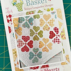 May include: A quilting pattern for a quilt called "Woven Hearts" by Margot Languedoc Designs. The pattern features a quilt with a white background and a design of woven hearts in shades of red, yellow, green, and blue. The pattern is available on the website www.thepatternbasket.com.