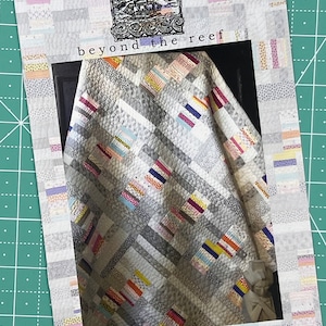 以下が含まれることがあります： モチによる「ビヨンド・ザ・リーフ」というキルトパターン。キルトは、パッチワークのデザインで、さまざまなカラフルな生地で作られています。キルトは暗い背景に表示されています。