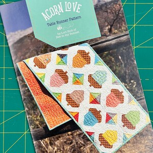 May include: A table runner pattern called "Acorn Love" by Lori Holt of Bee in My Bonnet. The pattern features a white background with colorful acorns in a repeating pattern. The pattern is shown on a table runner with a brown, orange, and blue color scheme.