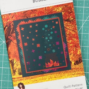 May include: Quilt pattern for a "Bubbling Brew" quilt, featuring a cauldron with stars and bubbles, set against a backdrop of autumn leaves. The pattern is by Sew A Story by Jennifer Long and the finished size is 74 inches by 86 inches.