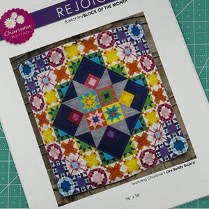 May include: A colorful quilt pattern called "Rejoice" is featured on a white paper with the text "8 Month/BLOCK OF THE MONTH" and "Featuring Charisma's Live Boldly Basics!" The quilt is 96 inches by 96 inches.