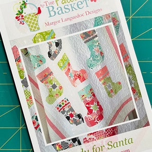 May include: A white and red quilt pattern with the title "Ready for Santa" and the website "www.thepatternbasket.com". The quilt features a patchwork design of Christmas stockings in various colours and patterns.