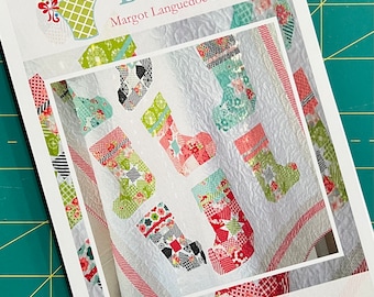 Może przedstawiać: Wzór kołdry w kolorze białym i czerwonym z tytułem "Ready for Santa" i stroną internetową "www.thepatternbasket.com". Kołdra ma patchworkowy wzór świątecznych skarpet w różnych kolorach i wzorach.