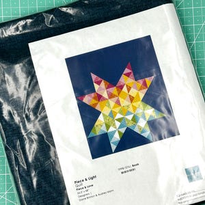 May include: A quilt kit in a plastic sleeve. The kit features a colorful star design on a navy blue background. The text on the packaging reads "Piece & Light Quilt Piece & Love 50.5 x 58".