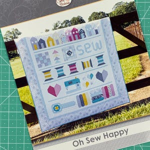 May include: A quilt pattern featuring a sewing theme with houses, spools of thread, buttons, and a sewing machine. The quilt is displayed on a wooden fence with the words "Oh Sew Happy" printed below.