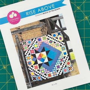 以下が含まれることがあります： 中央に星が描かれたカラフルなキルトブロックパターン。キルトブロックは、赤、青、黄、緑、黒など、さまざまな色の正方形と三角形で構成されています。キルトブロックは96インチ×96インチです。この画像は、カリスマ・ホートンによる「ライズ・アバブ」というキルトパターンブックからのものです。